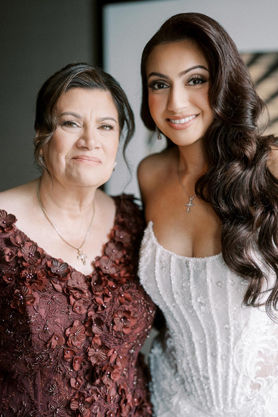 Two women smiling, one wearing a burgundy floral dress and the other in a white beaded gown, both with cross necklaces.