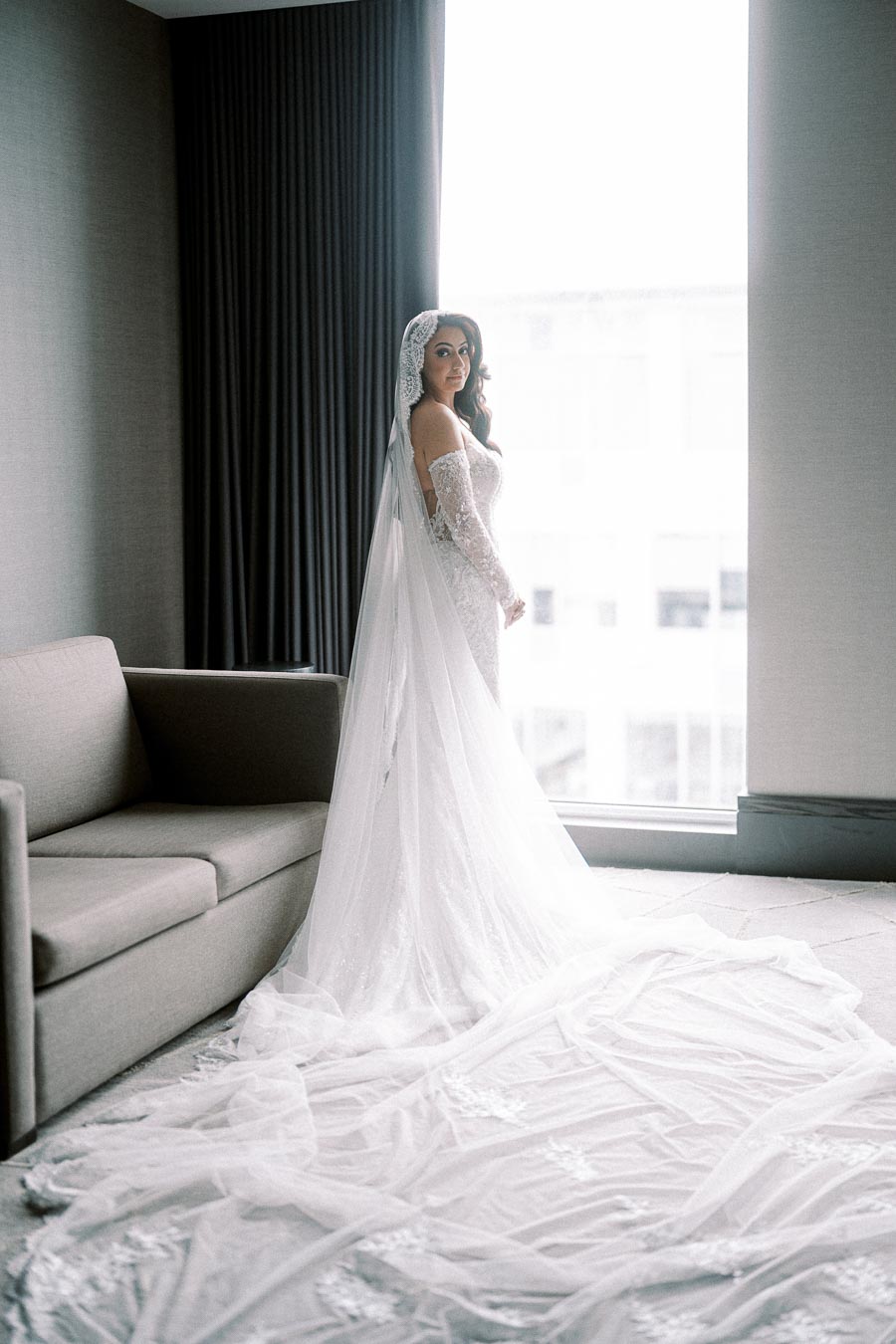 Bridal portrait featuring a woman in a stunning lace wedding dress with a long train, standing beside a large window in a