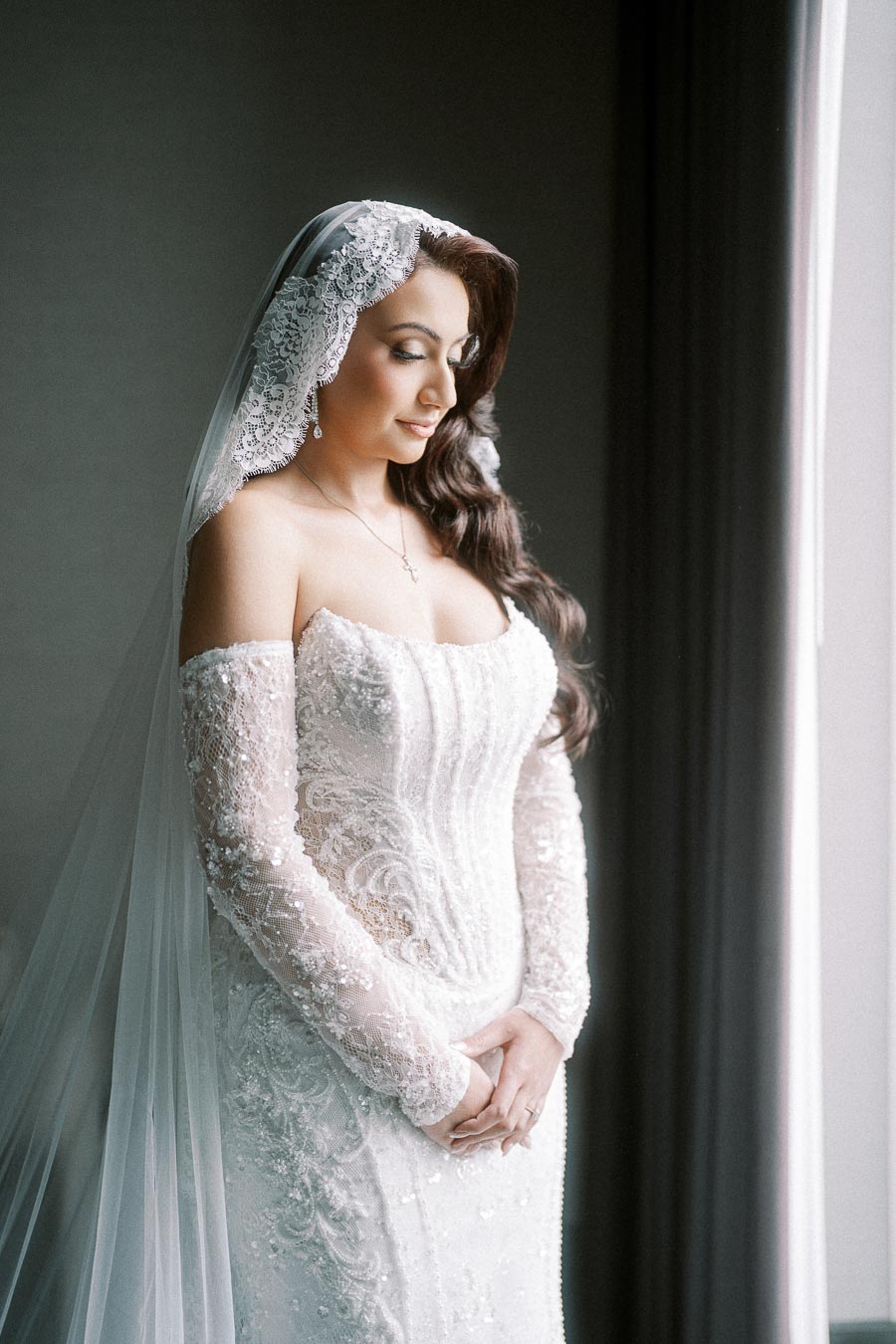Bride in an elegant lace wedding dress with off-the-shoulder sleeves, standing by a window with natural lighting.