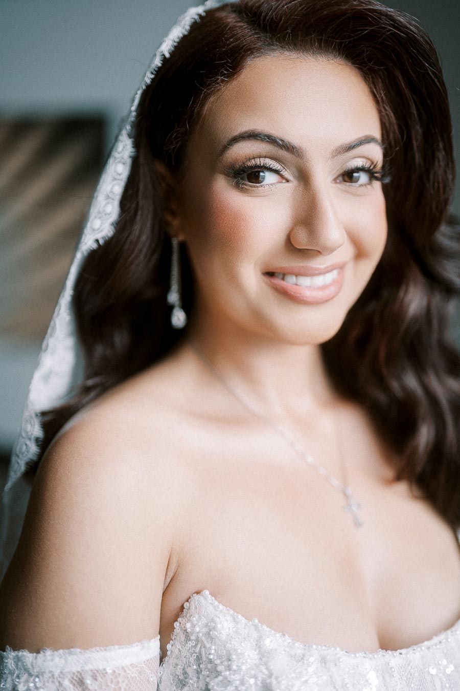 Elegant bride with wavy hair and lace veil smiling warmly, showcasing her bridal jewelry and exquisite wedding dress detail.