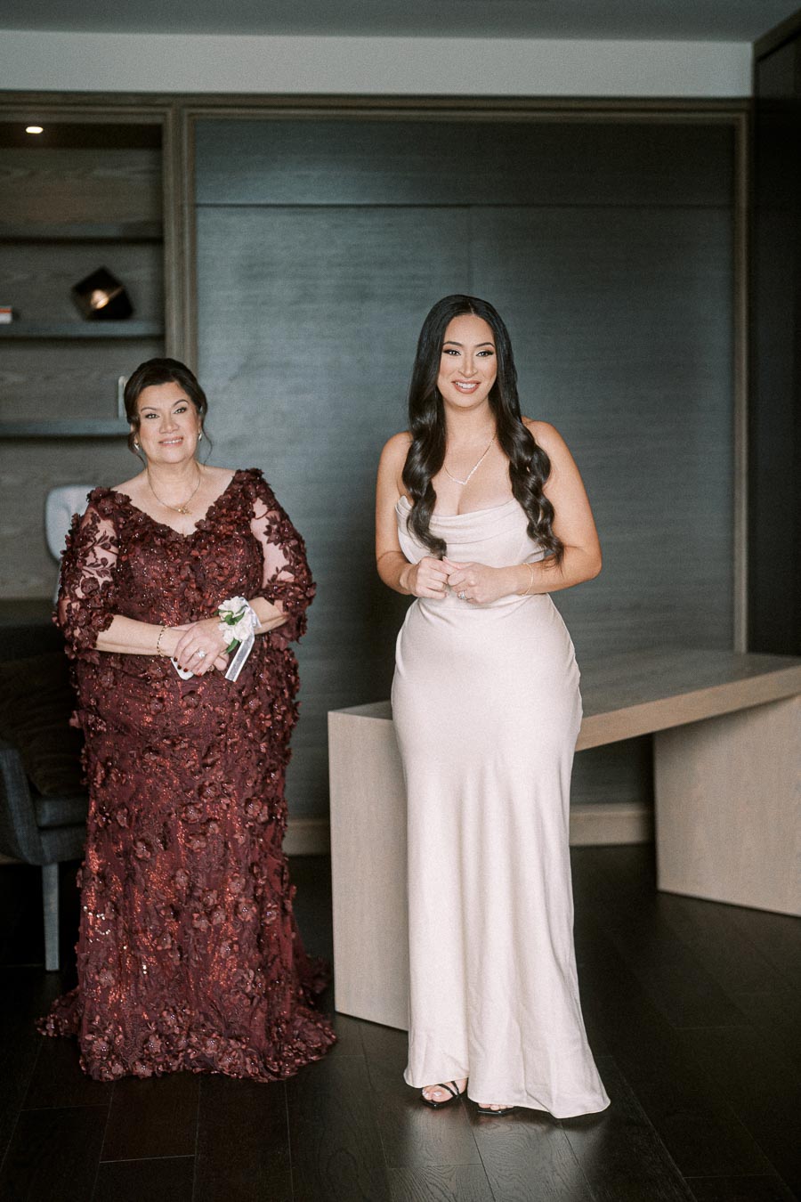 Two elegantly dressed women pose in a sophisticated indoor setting, one in a deep red floral gown and the other in a sleek