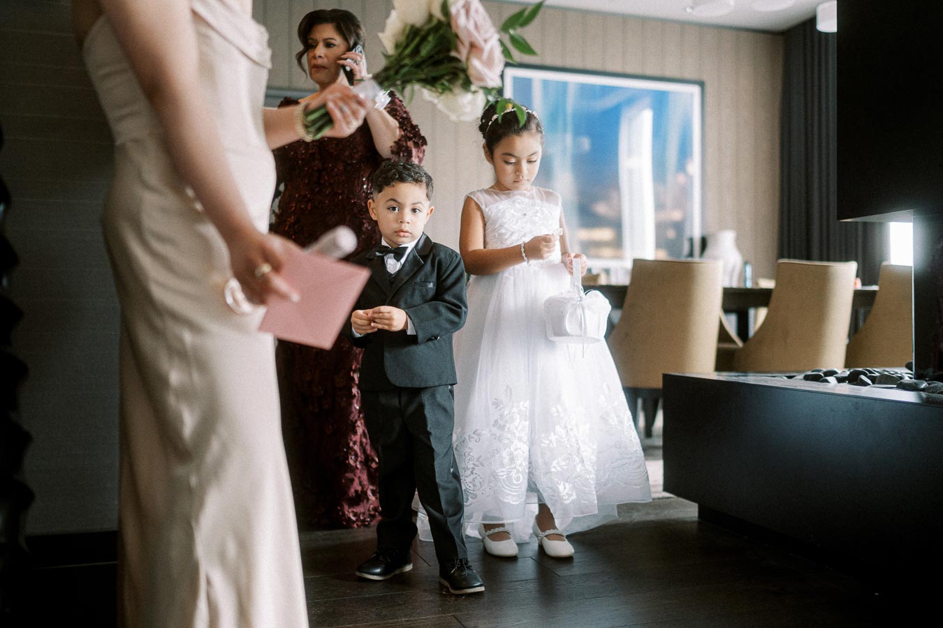 Children in formal attire preparing for a wedding, with a young boy in a tuxedo and a girl in a white dress holding a