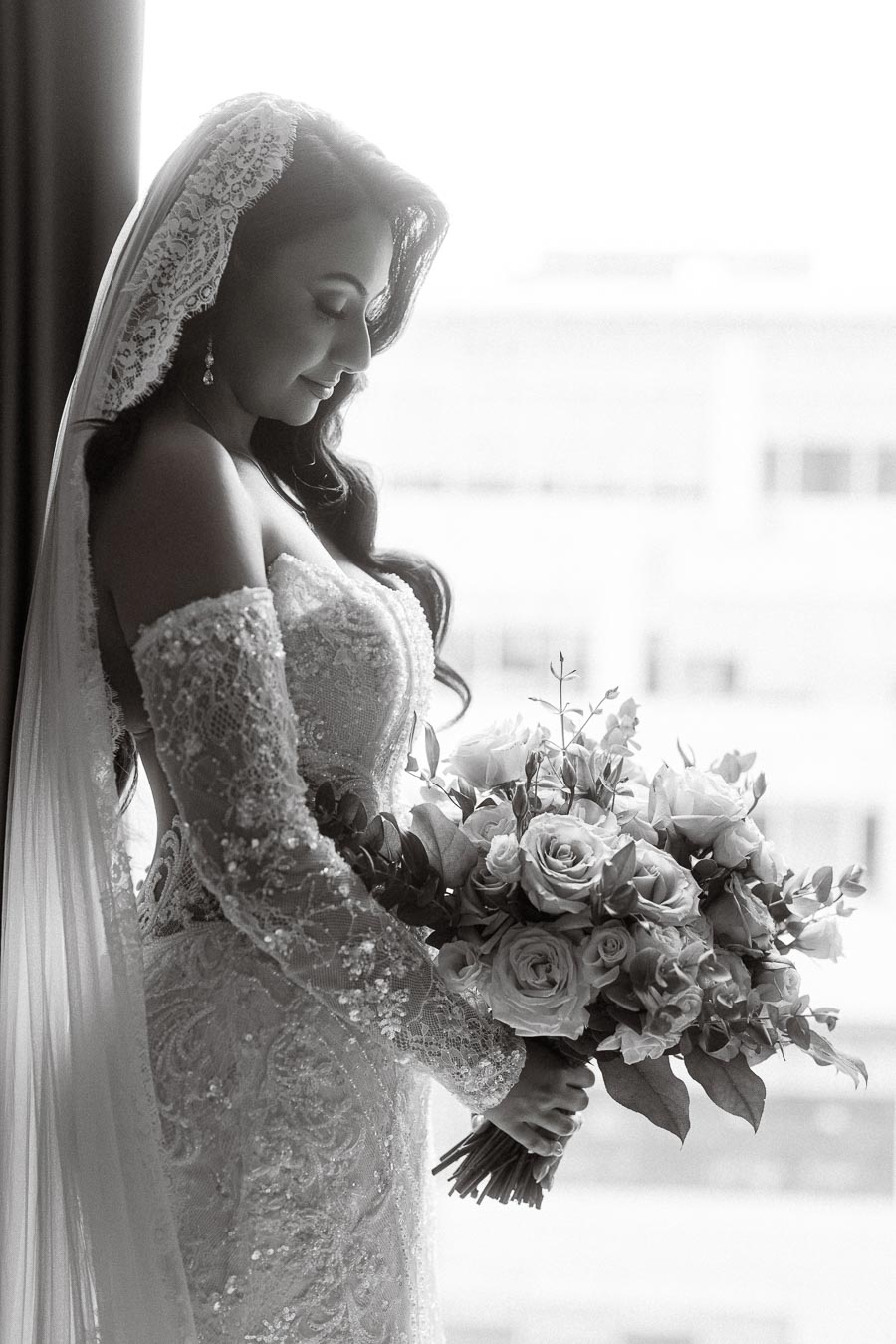 Elegant bride in a lace wedding gown holding a bouquet of roses, gazing down, with a soft, natural light background.