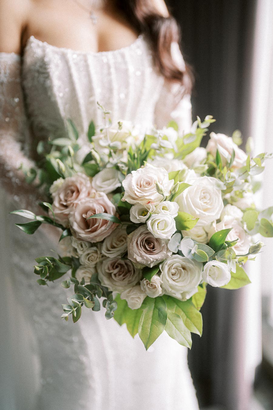 Elegant bridal bouquet featuring white and blush roses with lush green foliage, held by a bride in a shimmering white gown.