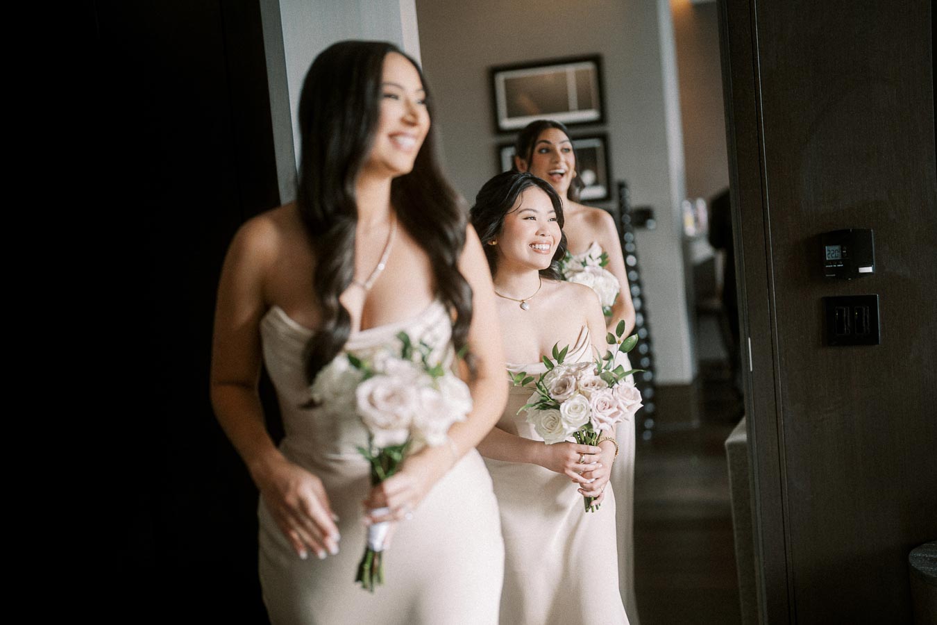Three smiling bridesmaids in elegant beige dresses hold delicate cream and pink rose bouquets, walking indoors with soft