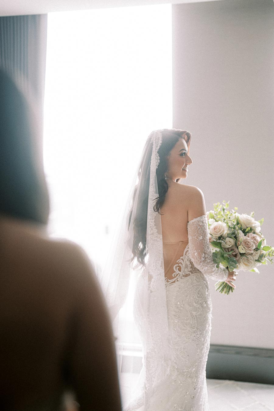 Bride in elegant lace wedding gown holding a bouquet of flowers, standing by a softly lit window.