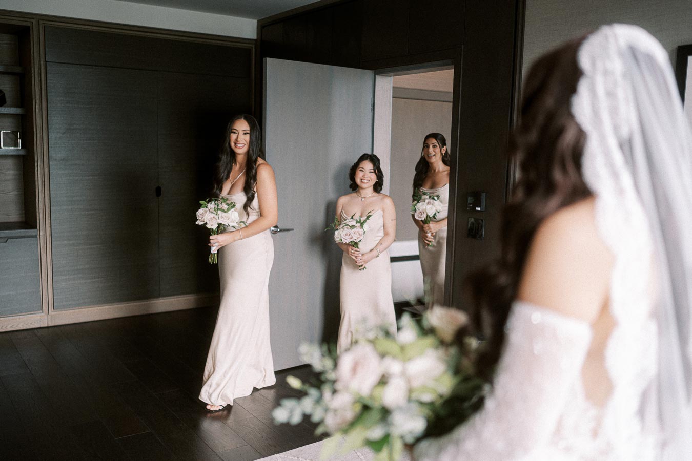 Bridesmaids in elegant dresses holding bouquets smile at the bride in a modern room setting.