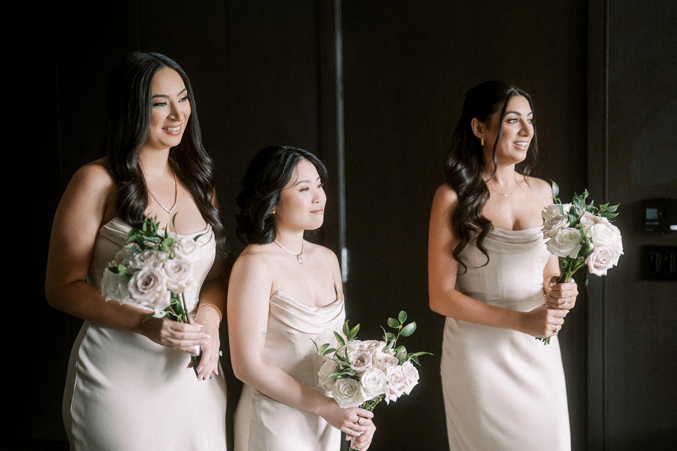 Three bridesmaids in elegant cream dresses hold bouquets of pink and white roses, standing together with smiles and