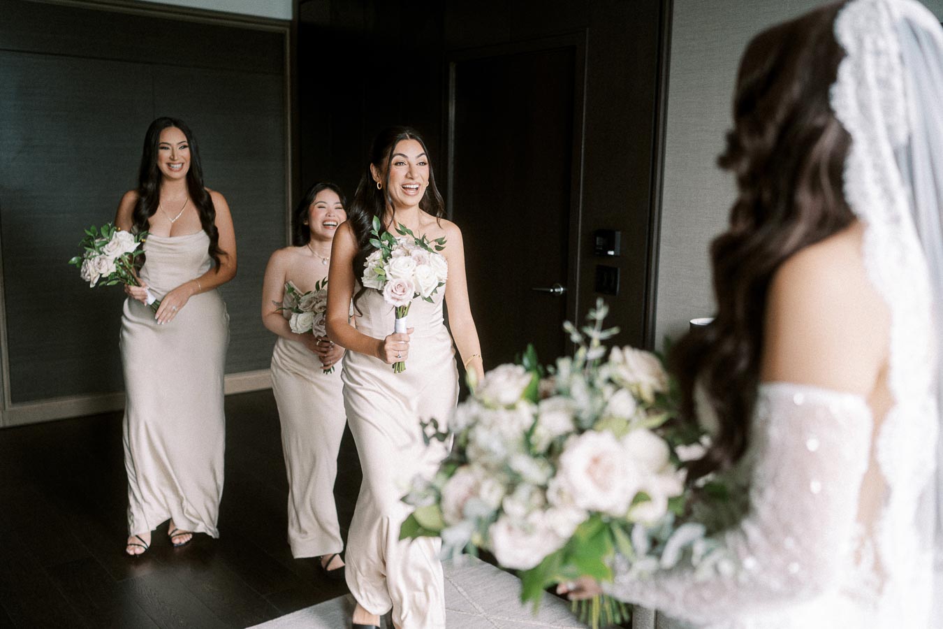Bridesmaids holding bouquets of white and pink flowers react joyfully upon seeing the bride in her wedding gown inside a
