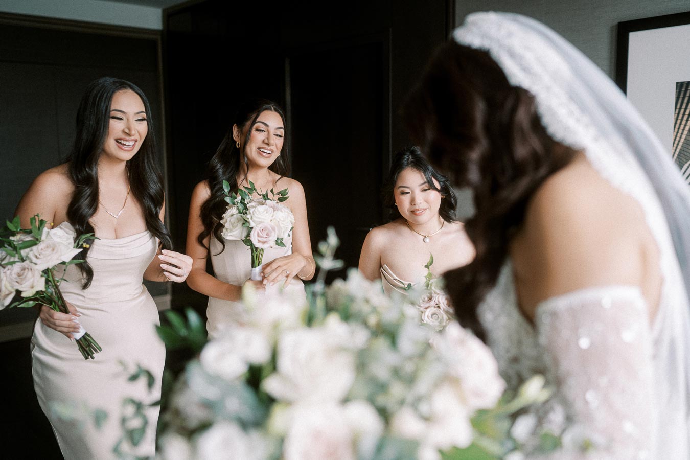 Bridesmaids in elegant dresses smiling and holding bouquets as they stand with the bride in a wedding preparation room.