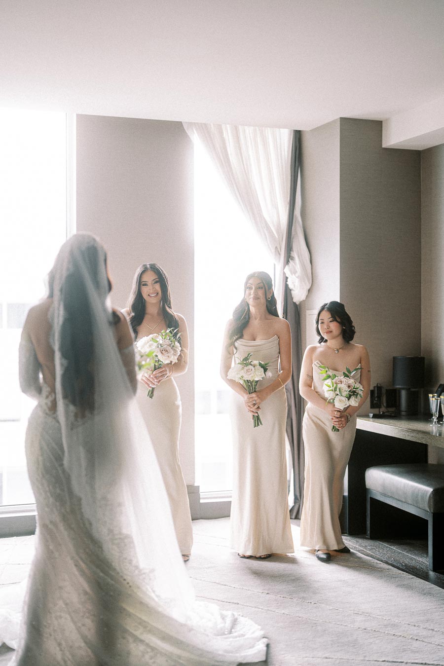 Bride with her back to the camera facing three smiling bridesmaids holding white bouquets in a bright, elegant room.