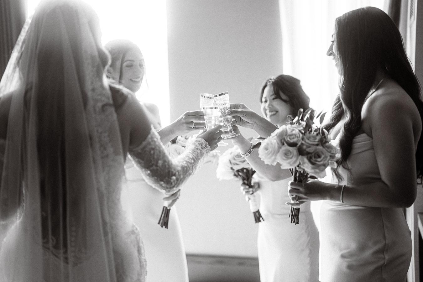 Black and white photo of four bridesmaids in elegant dresses toasting with champagne glasses, holding bouquets of