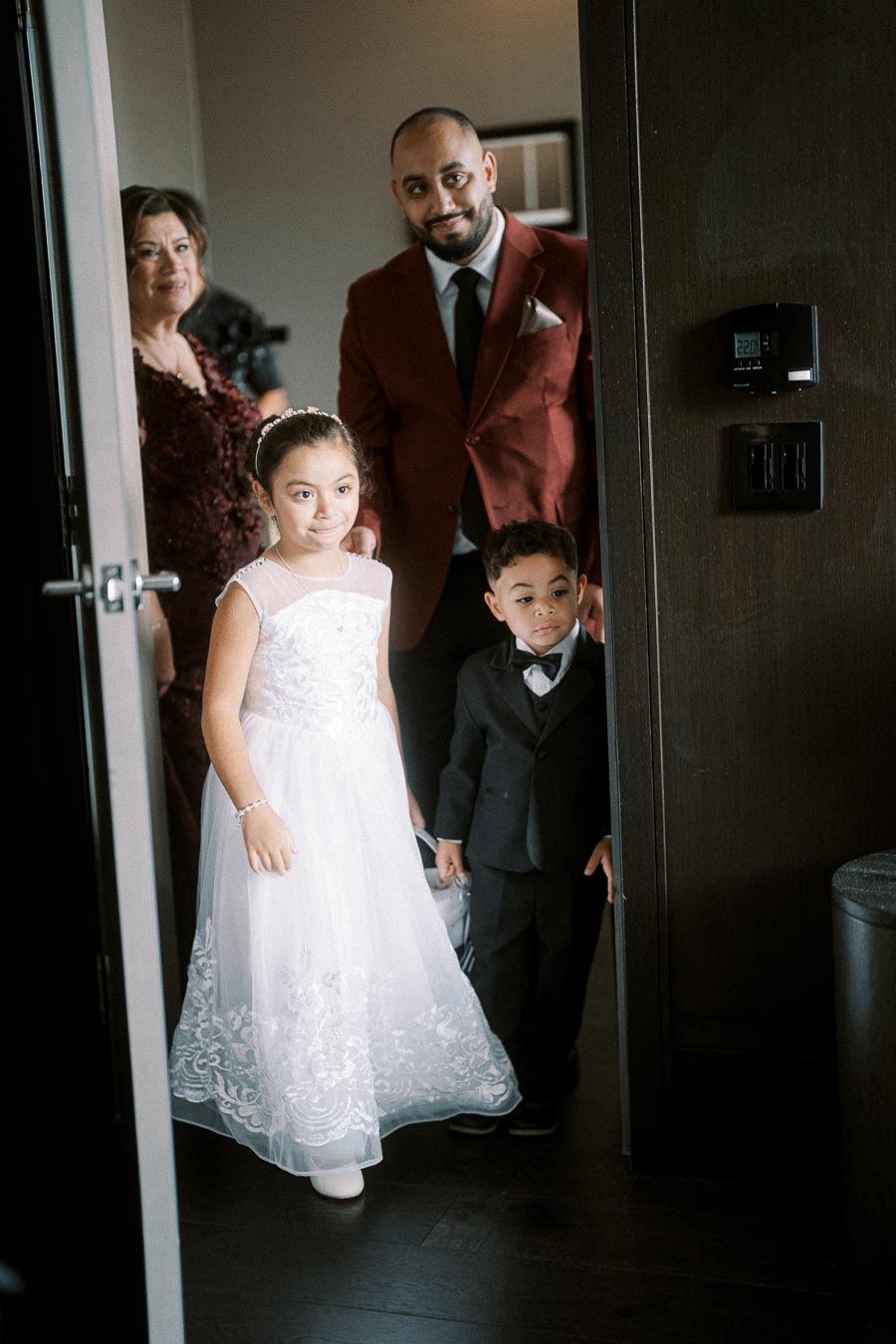 A young girl in a white dress and a boy in a suit stand in a doorway, accompanied by two adults, dressed for a formal event