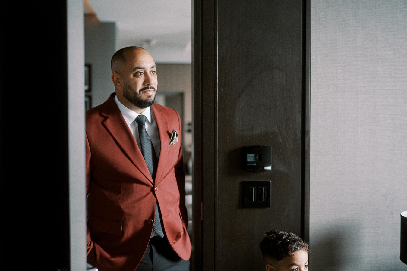 Man in a dark red suit and black tie standing by a modern interior doorway, with a child partially visible in the foreground.