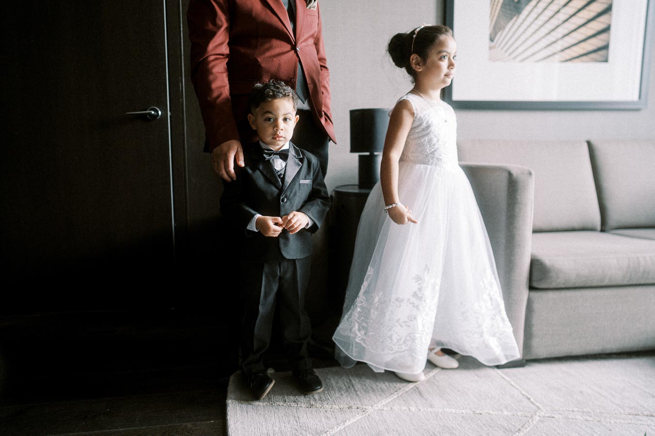 Two young children dressed in formal attire stand in a modern room; the boy is wearing a black suit with a bow tie, and the