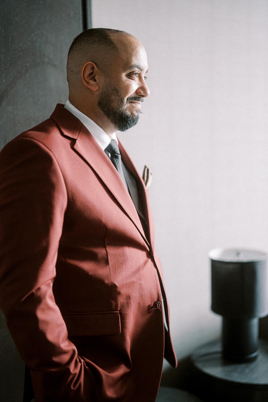 A man in a stylish red suit stands confidently, with light highlighting the texture of his outfit. He has a neat beard and