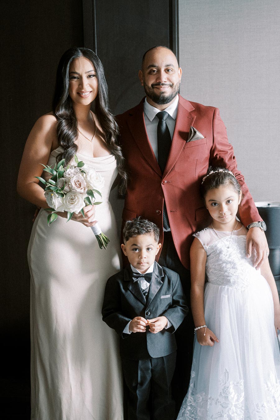 A happy family posing together in formal attire; the woman holds a bouquet and wears a white dress, while the man is dressed