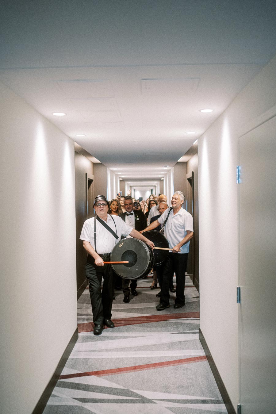 A lively group of musicians with drums leading a parade down a hotel hallway, followed by a smiling crowd dressed for a