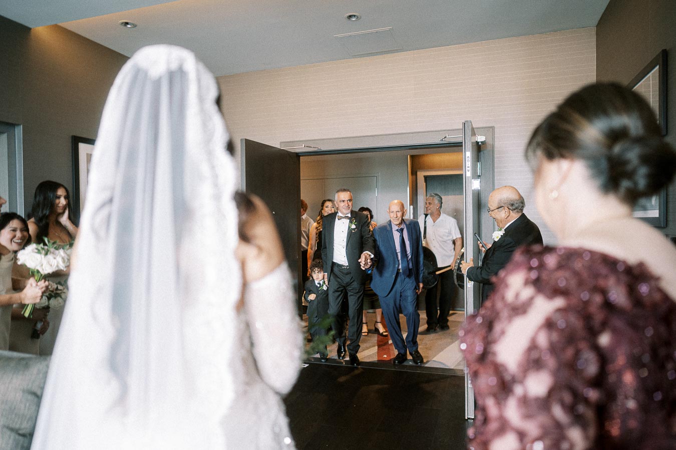 A bride in a white gown is facing a group of people entering a room during a wedding ceremony. The guests are dressed