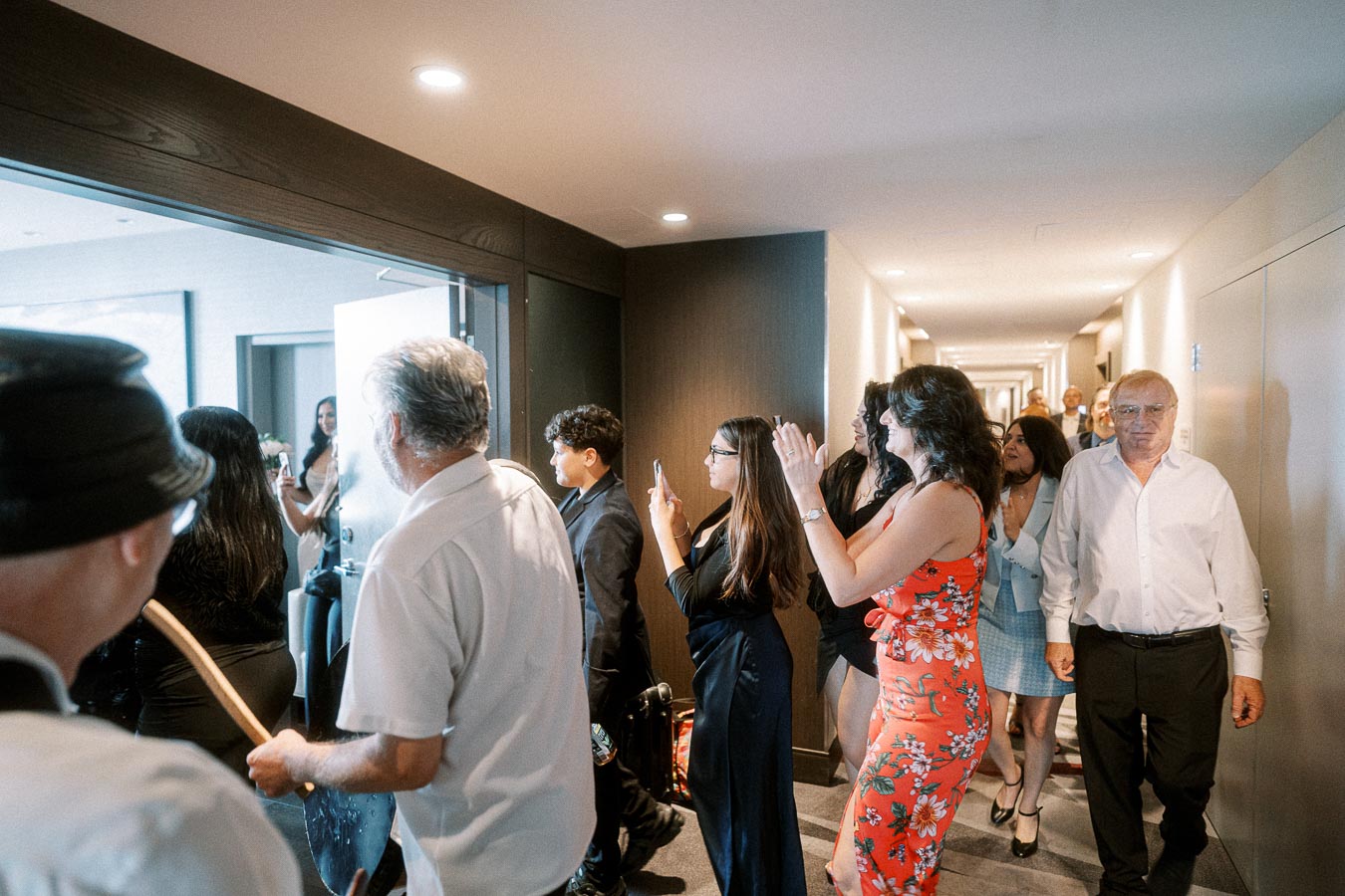 Group of people dressed formally, gathered in a hotel corridor, celebrating with smiles and phones as a door opens to reveal
