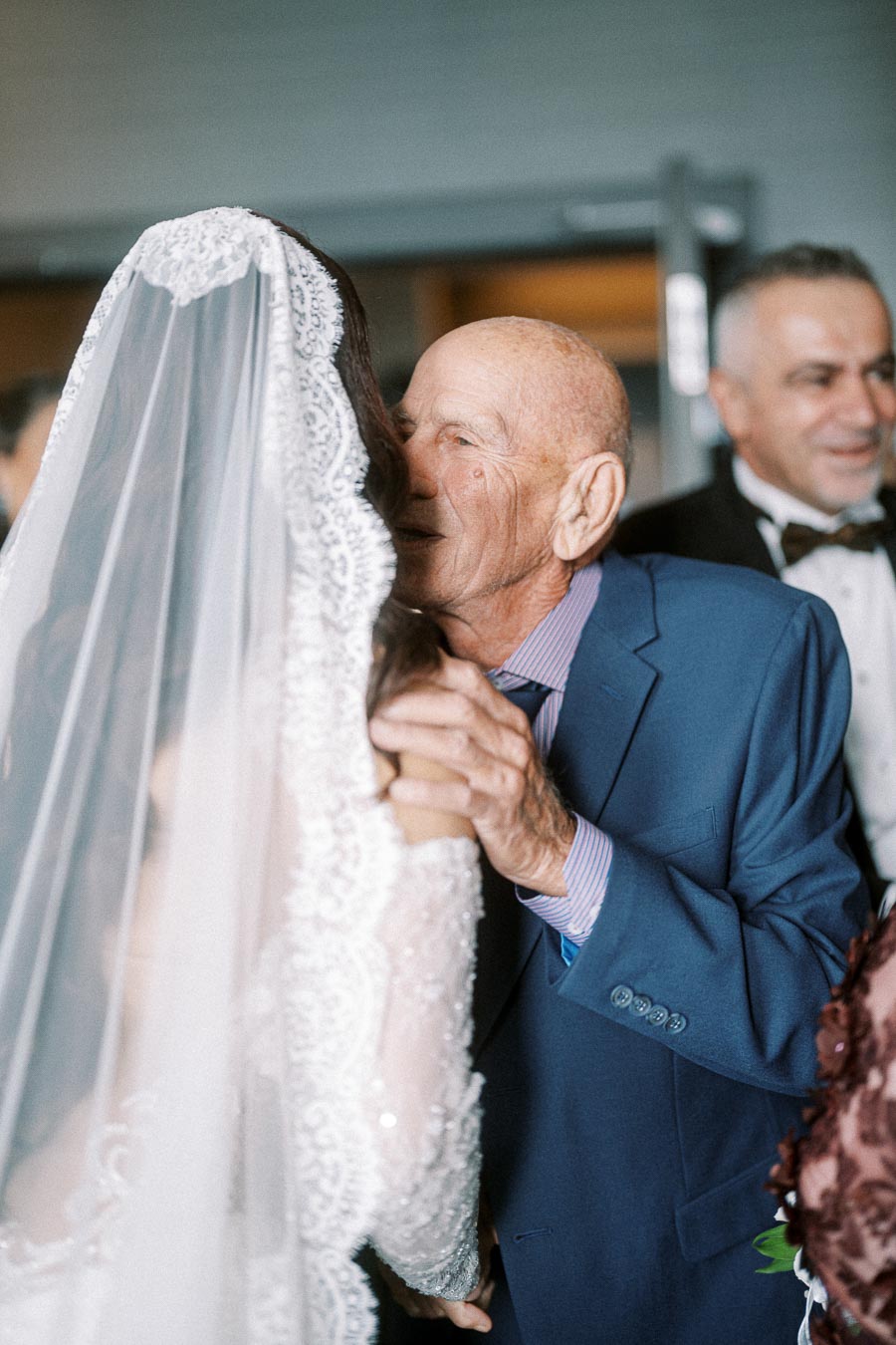 Emotional moment between a bride in a lace wedding dress and an elderly man in a blue suit, captured at a wedding ceremony.