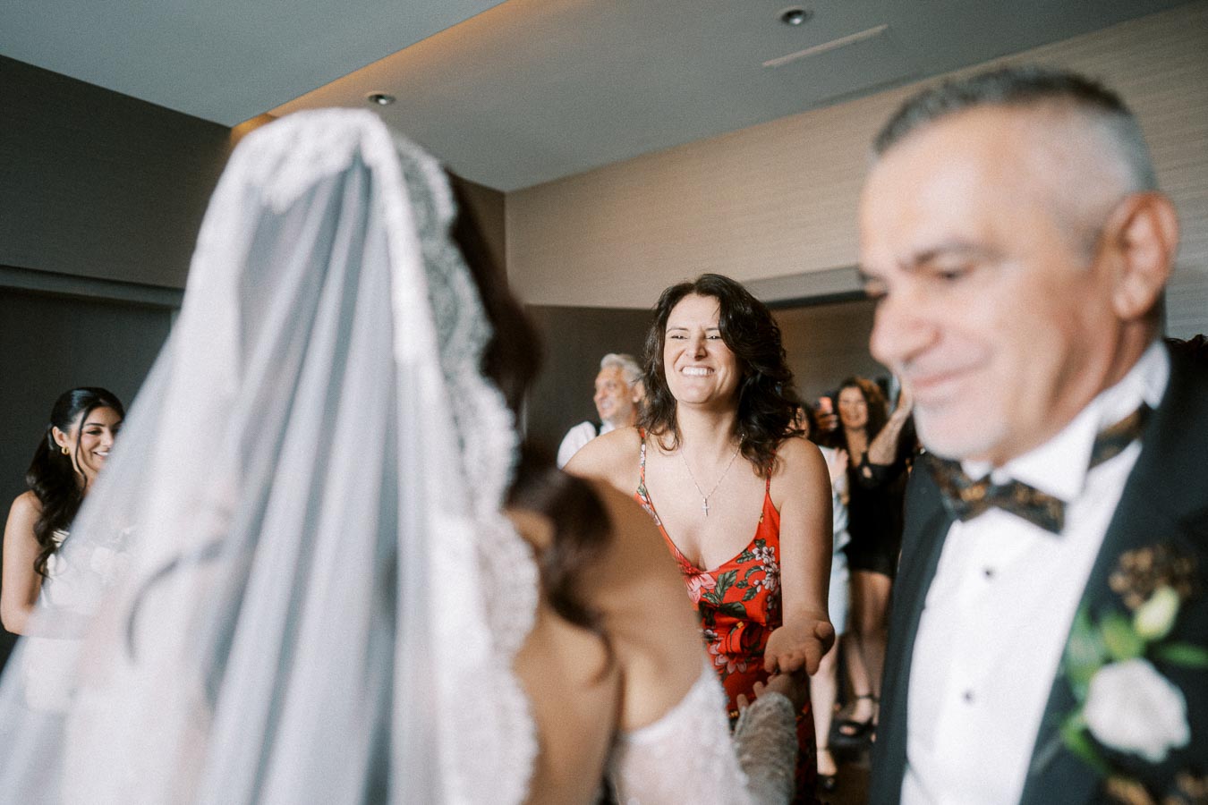 A bride in a lace wedding dress greets a smiling woman in a floral dress at a wedding reception. A man in a tuxedo stands