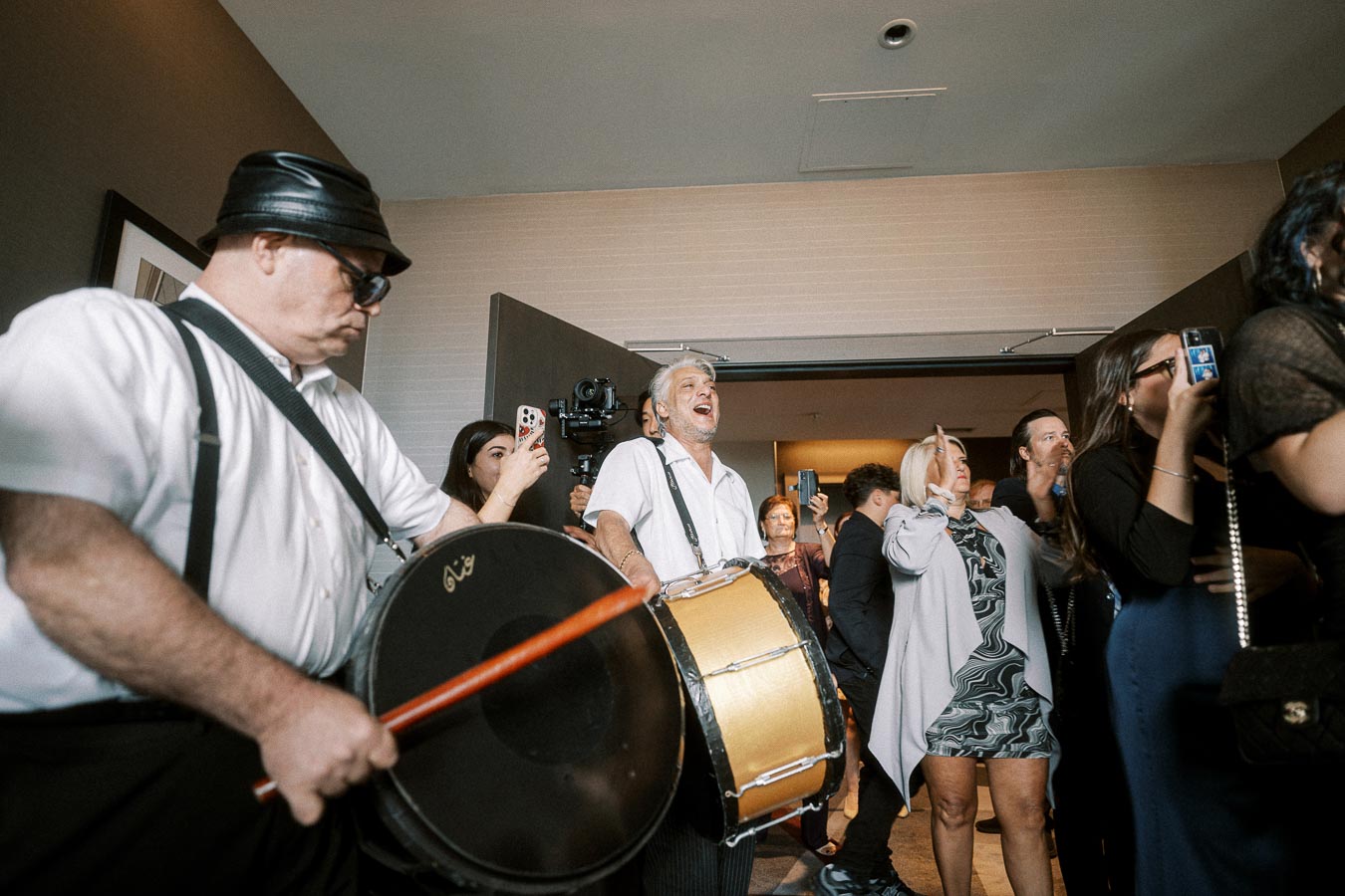 Group enjoying live drumming performance at indoor event, with onlookers capturing the moment with smartphones.