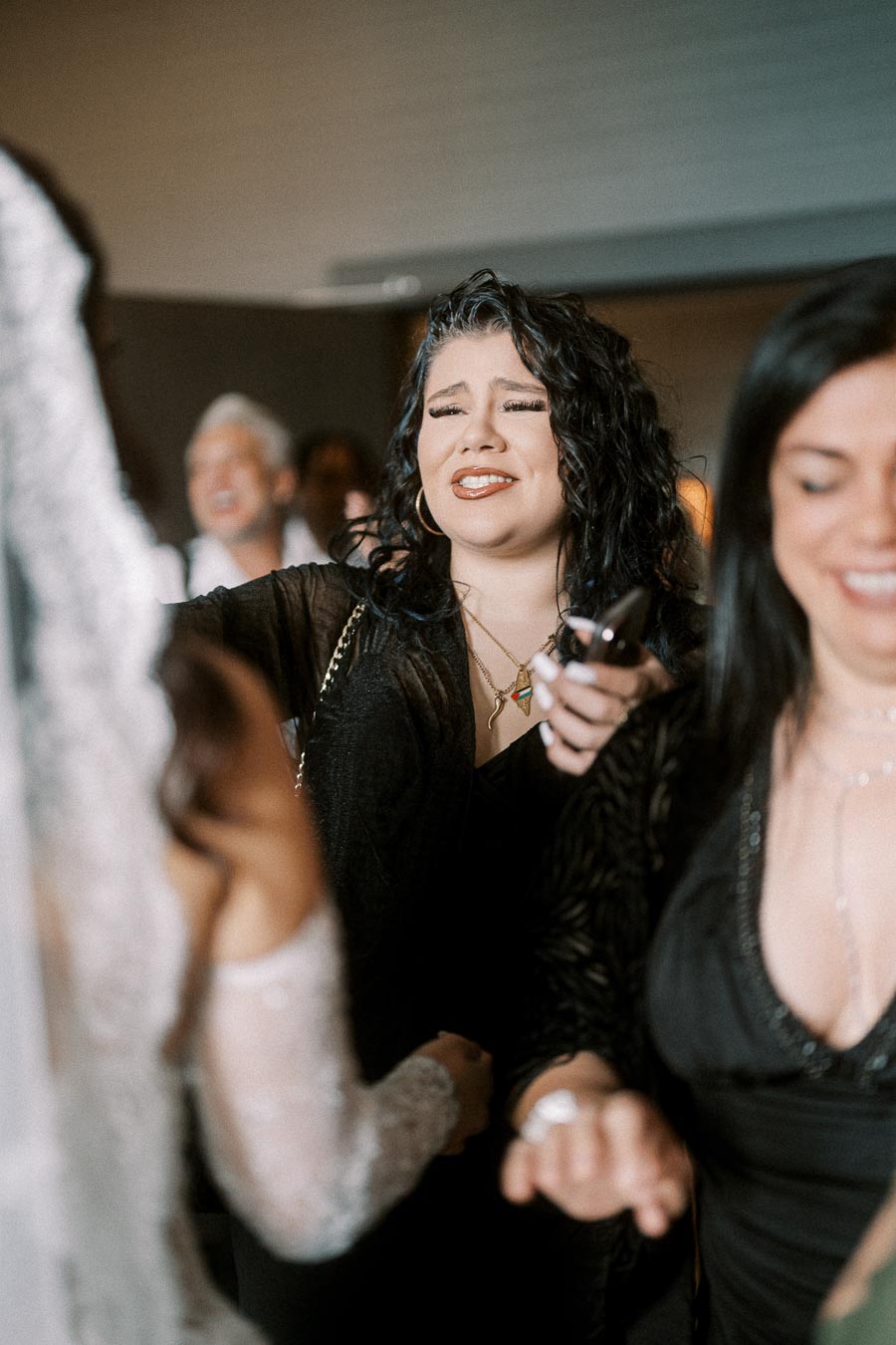 A woman with curly dark hair and a joyful expression, dressed in a black outfit, sings and enjoys a lively indoor event,