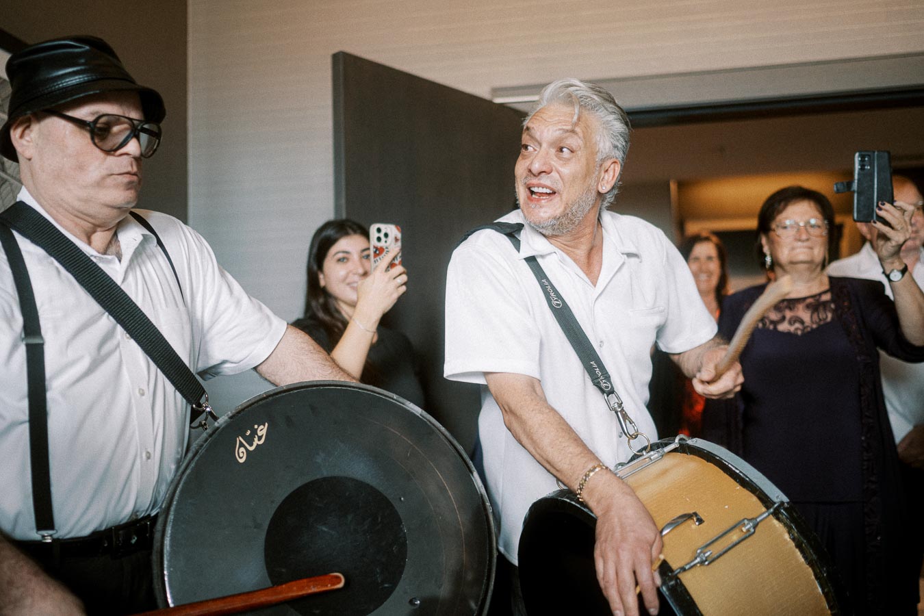 Two men playing drums at a lively indoor gathering, with enthusiastic guests capturing the moment on their phones.