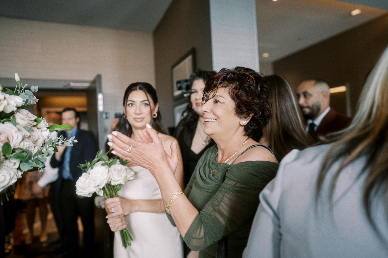 A joyful group of people celebrates at a wedding reception, featuring a smiling woman in a green dress clapping, surrounded