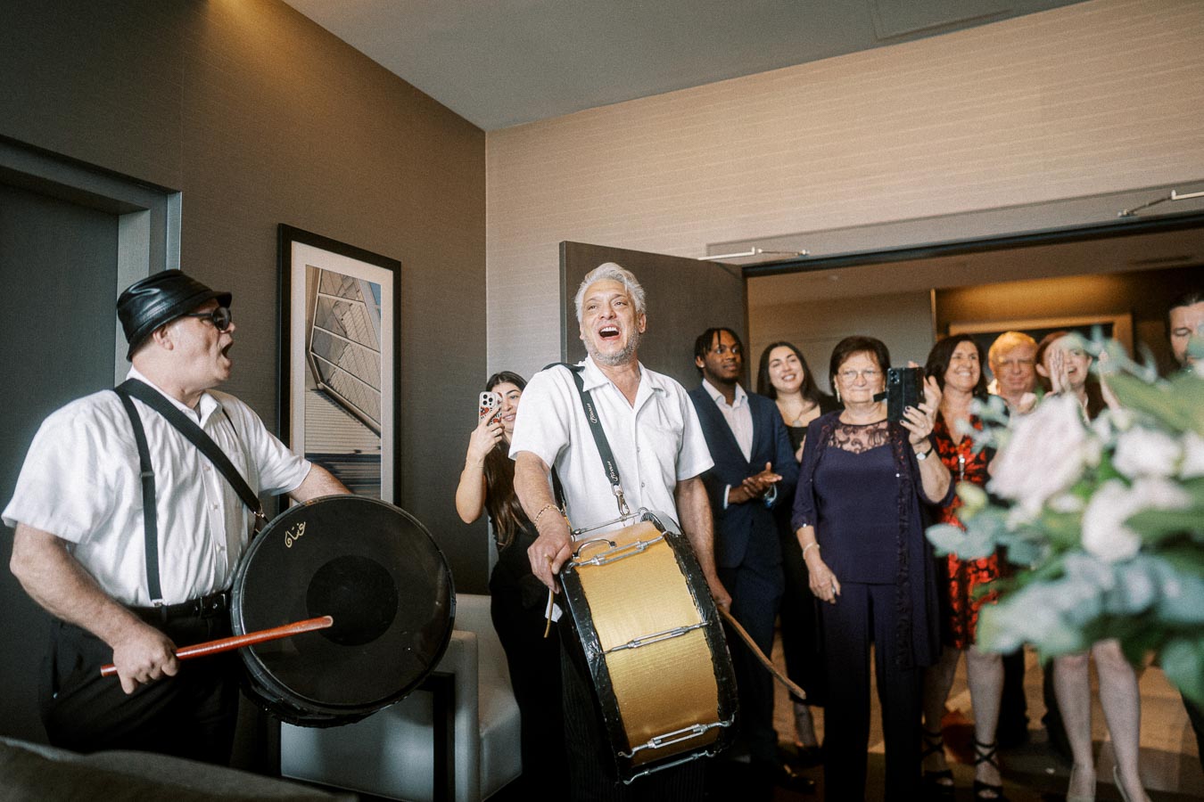 A lively group of people enjoying a celebration indoors, featuring two musicians playing drums, with others clapping and