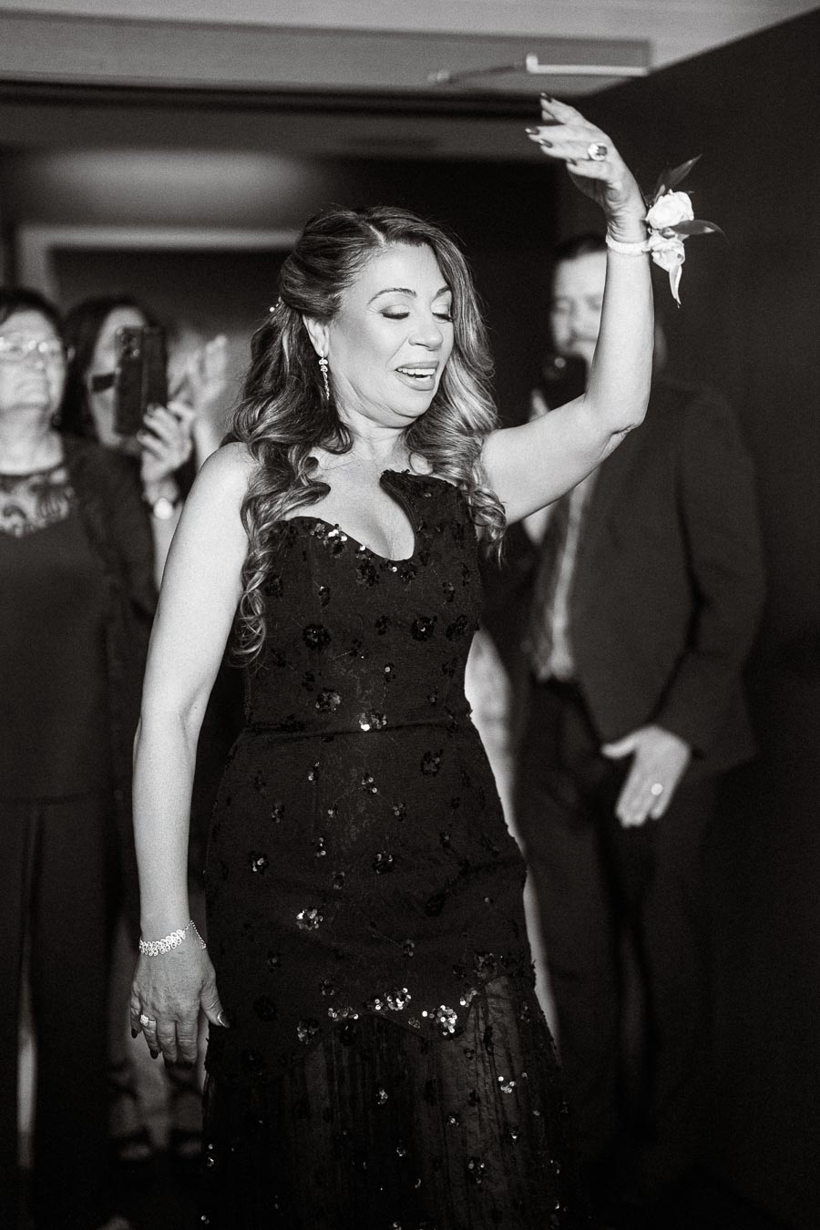 A woman in an elegant black dress with sequin details raises her hand at an event. She has wavy hair and is wearing a