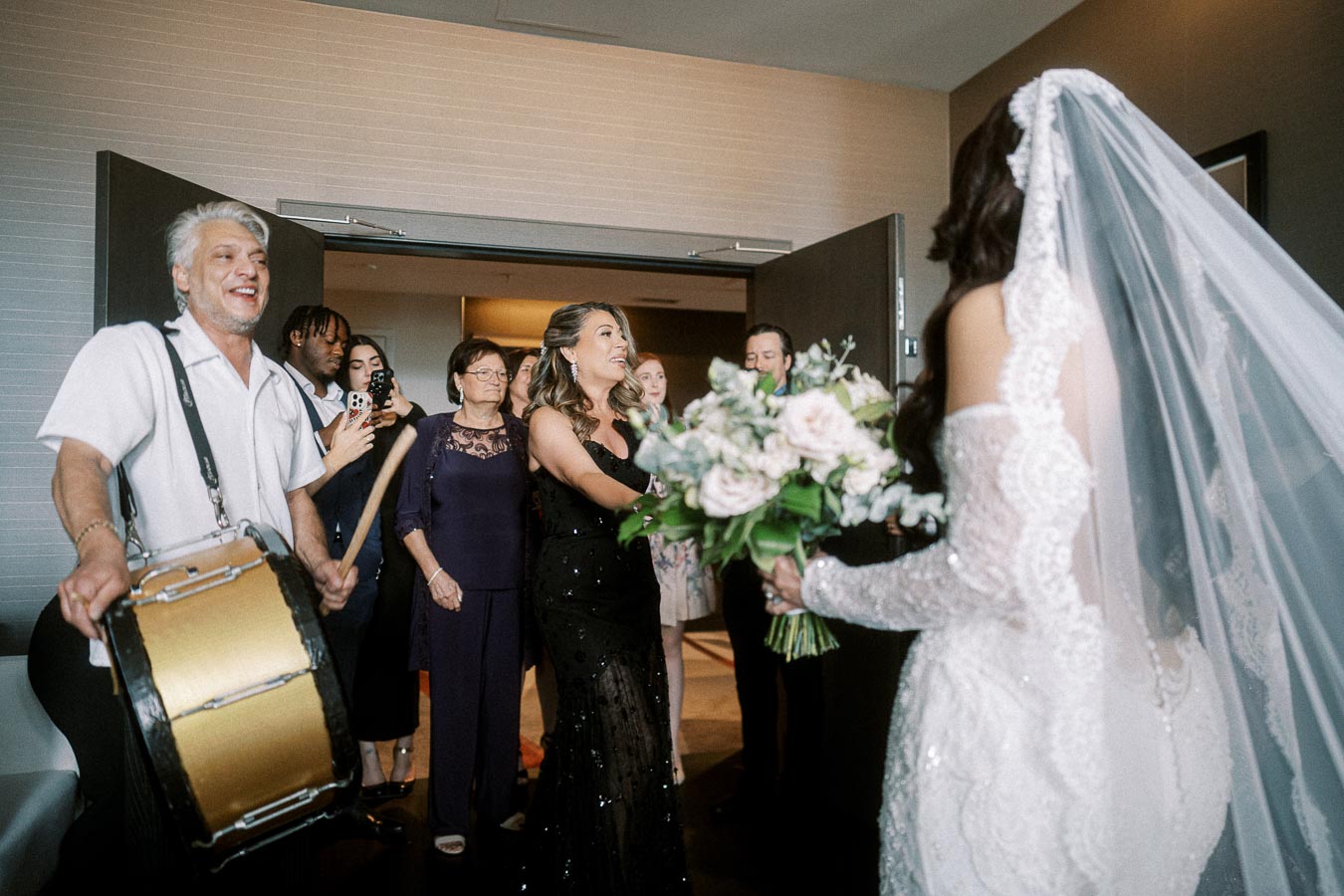 Wedding celebration with a bride in a lace gown holding a bouquet, surrounded by smiling guests, and a drummer playing in