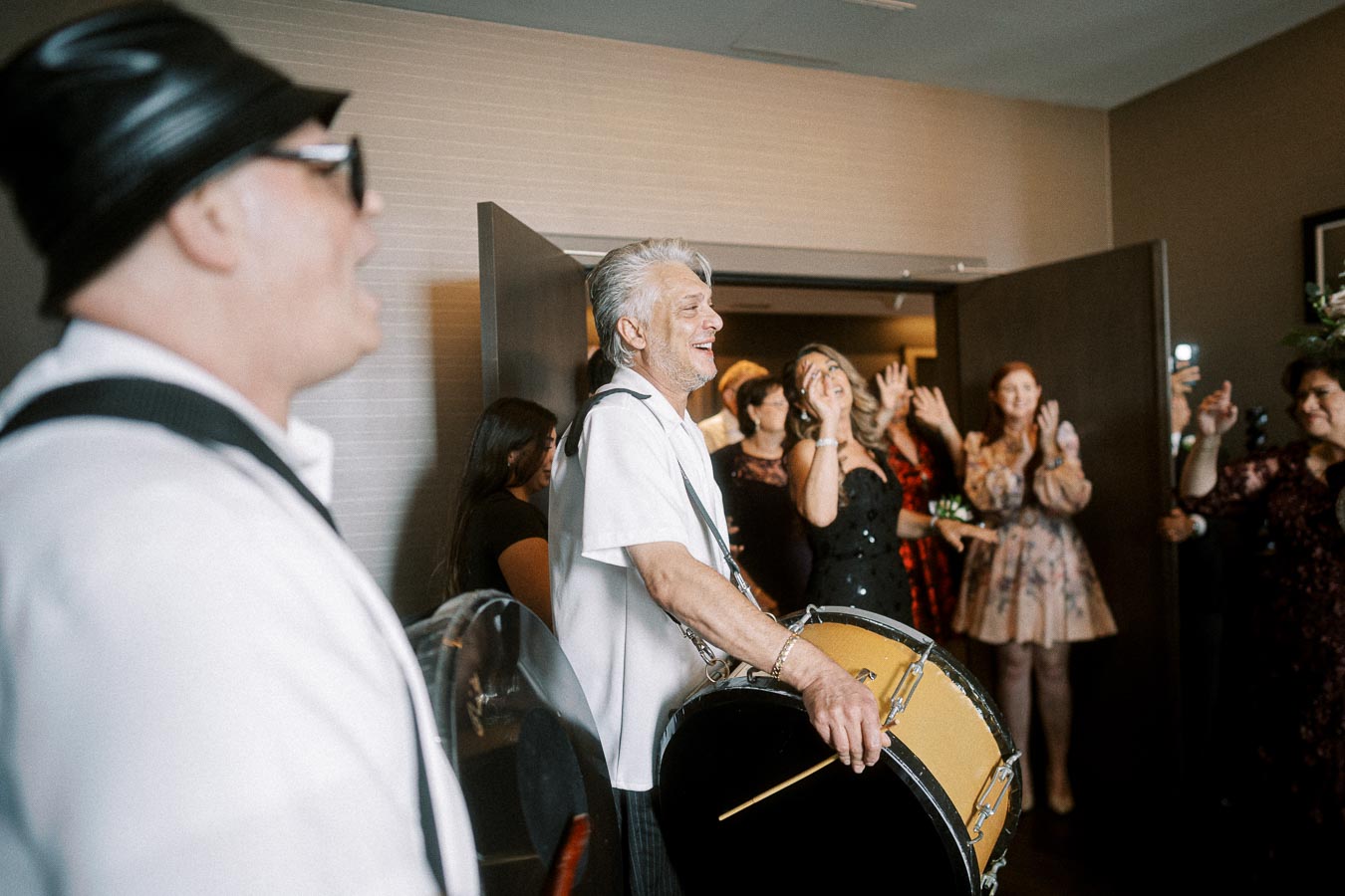 Man playing drum at lively indoor event with guests dancing and enjoying music.