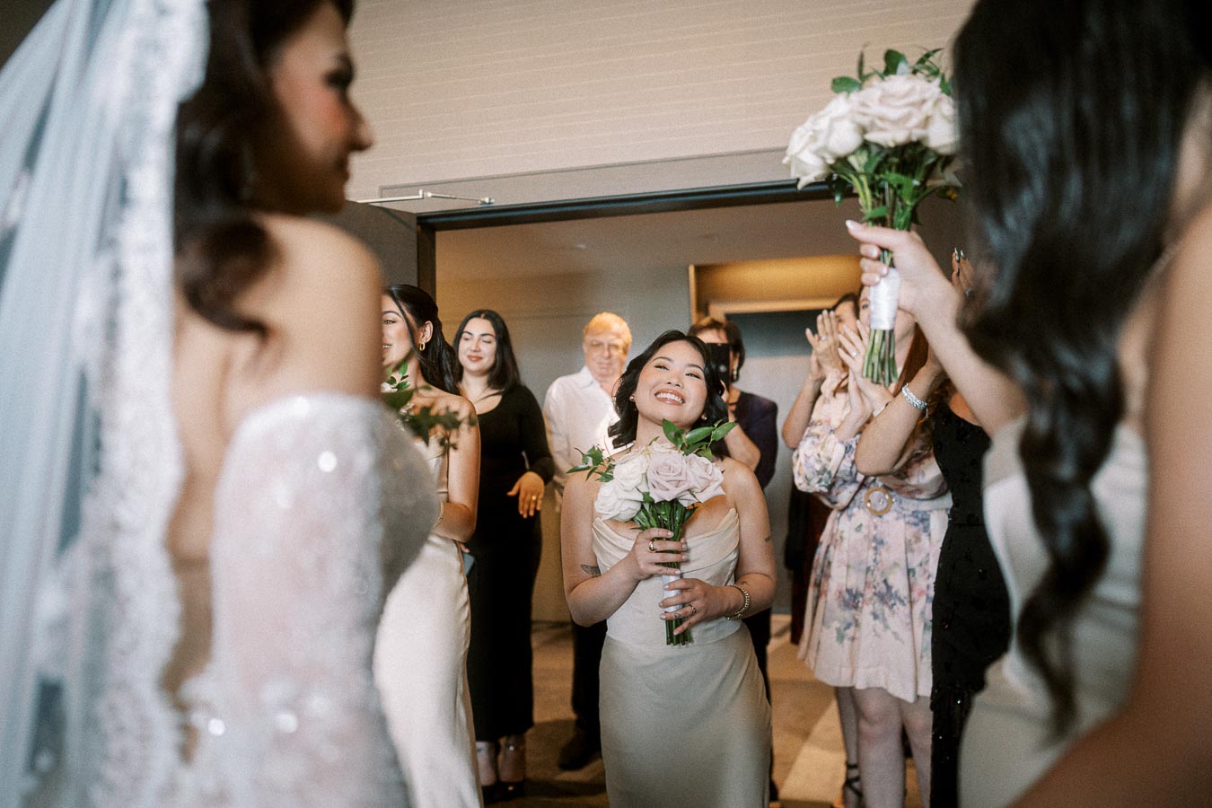A joyful bridesmaid holding a bouquet of roses, surrounded by other smiling guests at a wedding celebration indoors.