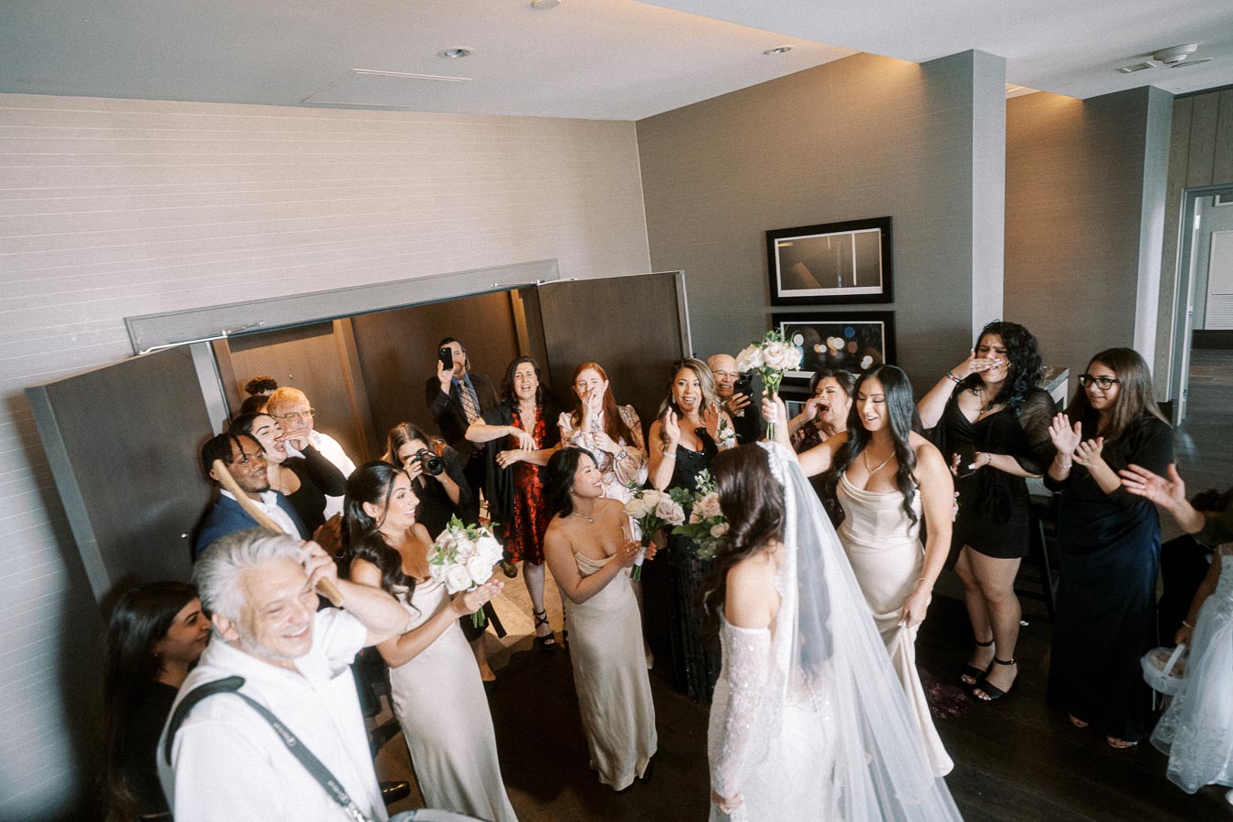 A bride in a wedding dress is surrounded by her excited bridesmaids and friends in a stylish room, capturing a joyful