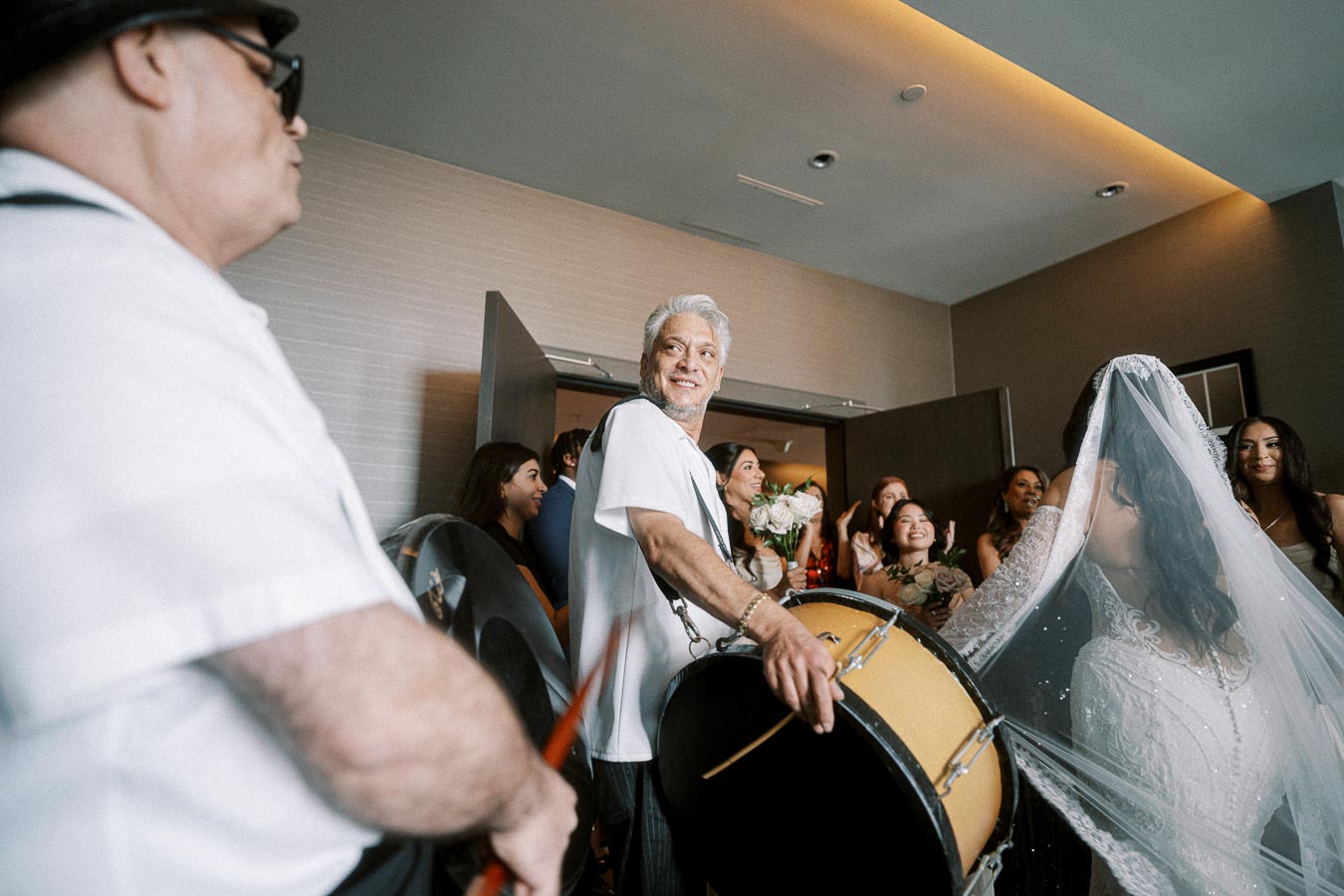 A joyful wedding celebration indoors featuring a smiling drummer wearing a white shirt, playing a large drum. A bride in a