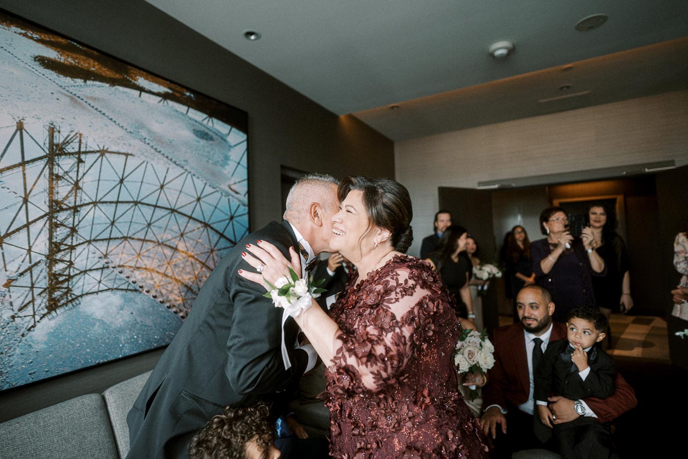 Elderly couple embracing warmly at a family gathering, surrounded by smiling guests, in a stylish room with modern art on