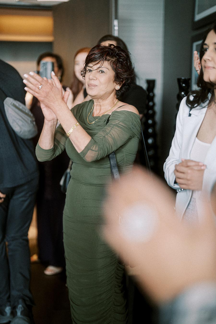 Elegant woman in a green dress clapping during a social gathering, surrounded by people celebrating indoors.