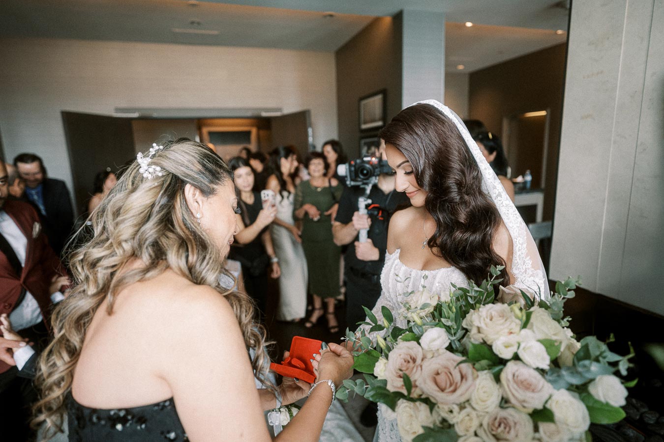 Bride receives gift during intimate pre-wedding gathering with family and friends, holding a bouquet of roses and greenery.