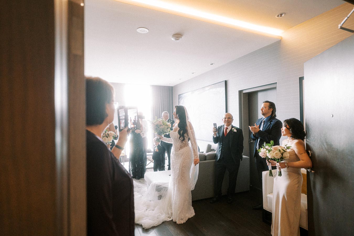 A bride in a lace wedding dress holding a bouquet enters a room filled with family and friends capturing the moment on their