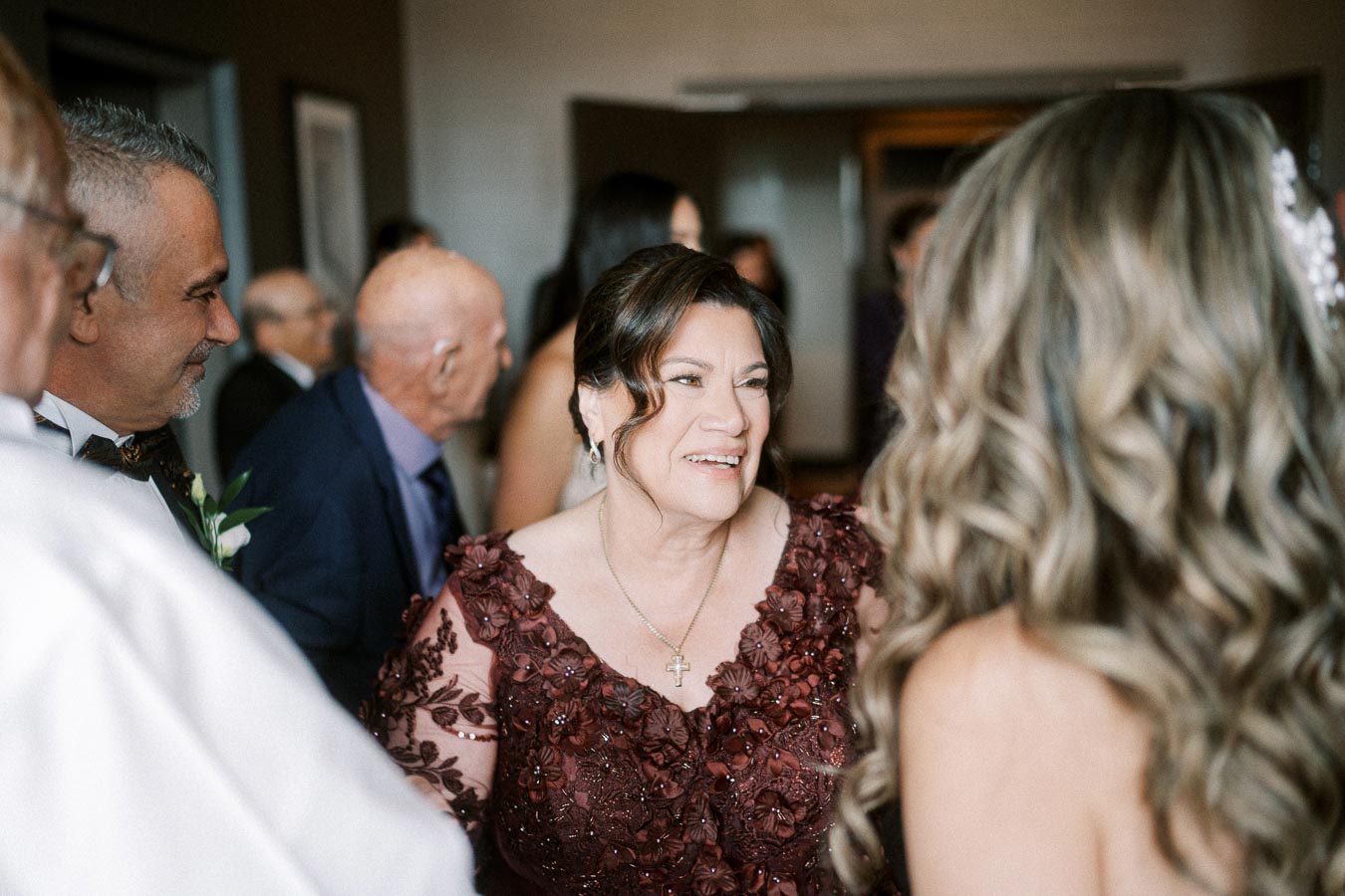 Group of people socializing at an indoor event, with a woman in a detailed maroon dress smiling and engaging in conversation.