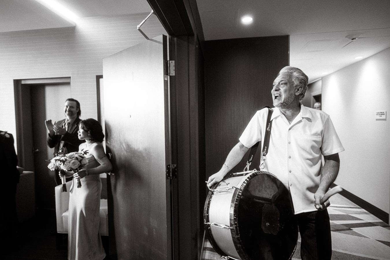 Black and white photo of a joyful man playing a traditional drum in a hallway, with a smiling bride holding a bouquet and an