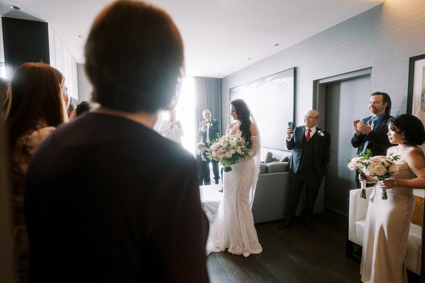 Bride in elegant white gown enters a room filled with joyful guests, carrying a bouquet of roses, as a photographer captures