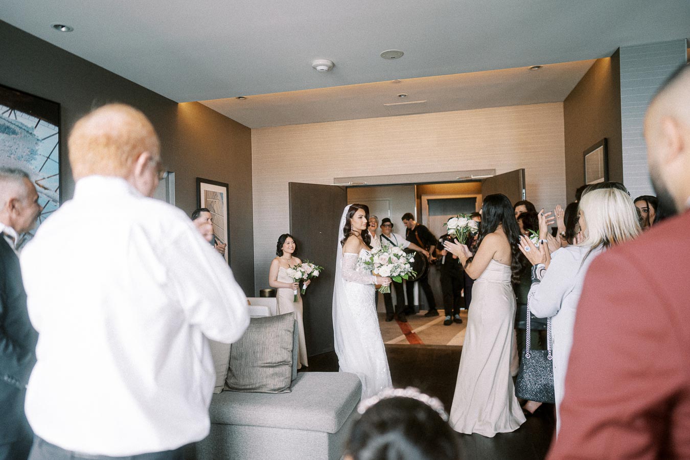 A bride in a white lace gown and veil enters a room filled with family and friends, who are smiling and applauding. She