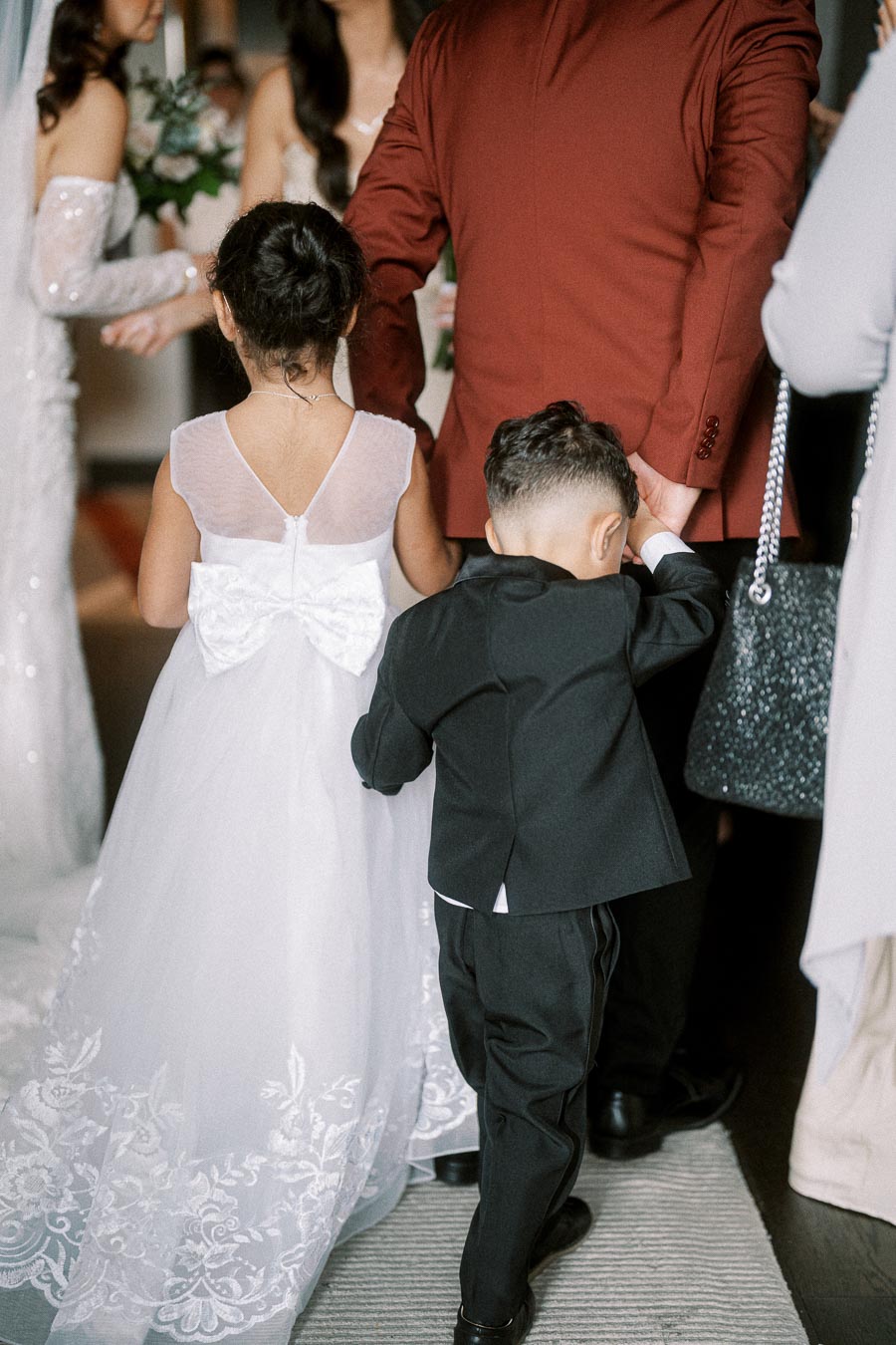 A young girl in a white dress with a large bow and a boy in a black suit are walking together at a wedding ceremony,