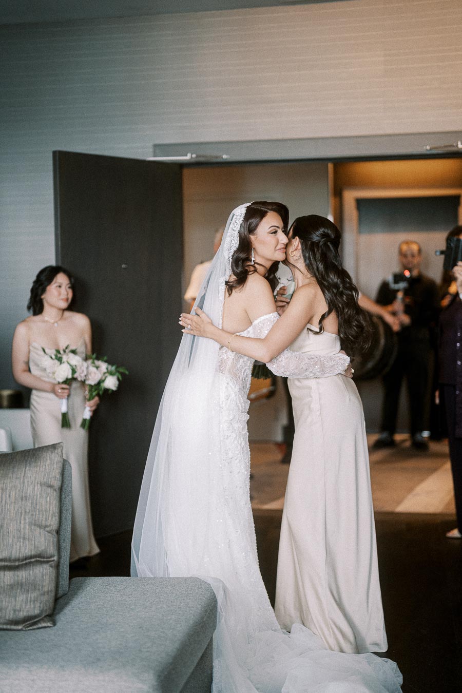 Bride in elegant white gown embraces bridesmaid in beige dress during wedding preparation, with another bridesmaid holding a