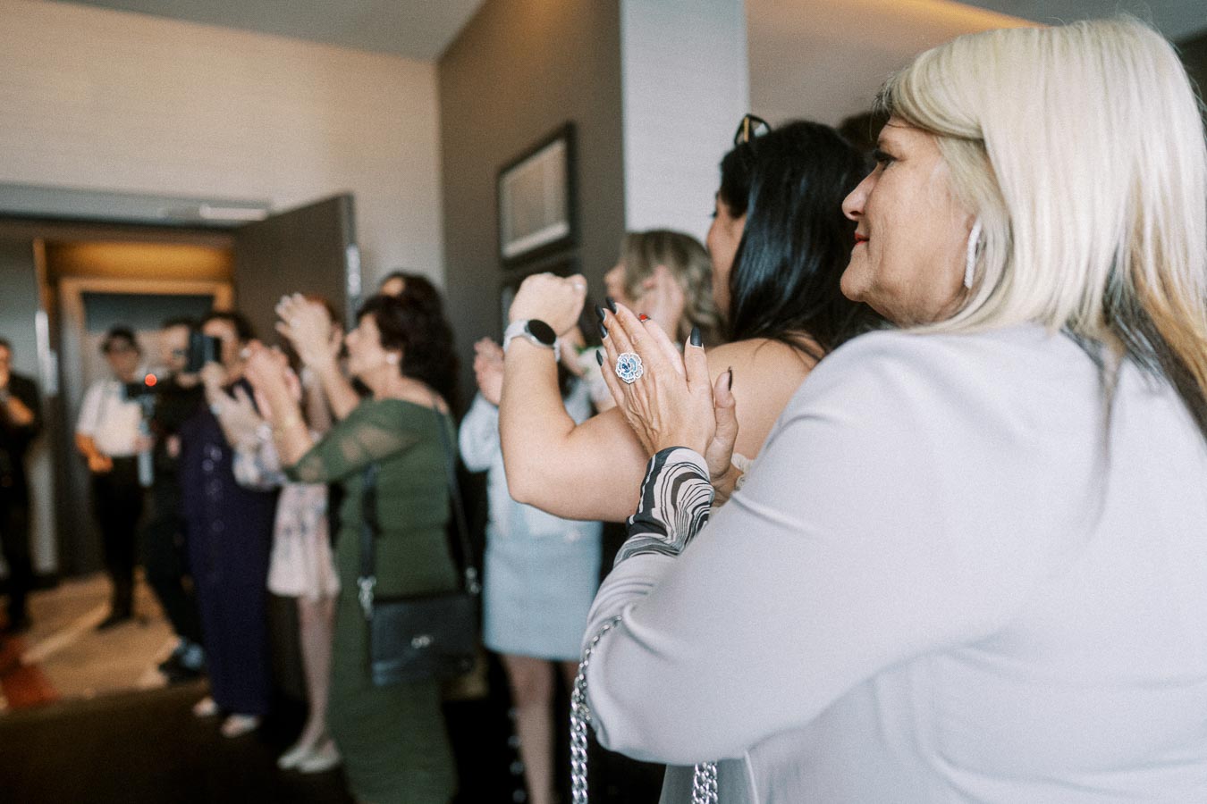 A group of people applauding in a conference room, with a woman in the foreground wearing a white top and a large ring,