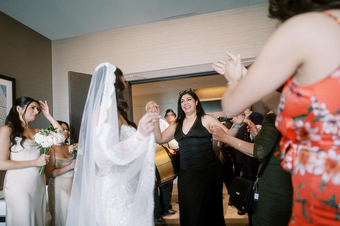 A joyful bride in a white dress is surrounded by smiling guests celebrating at a wedding. Bridesmaids hold bouquets, and