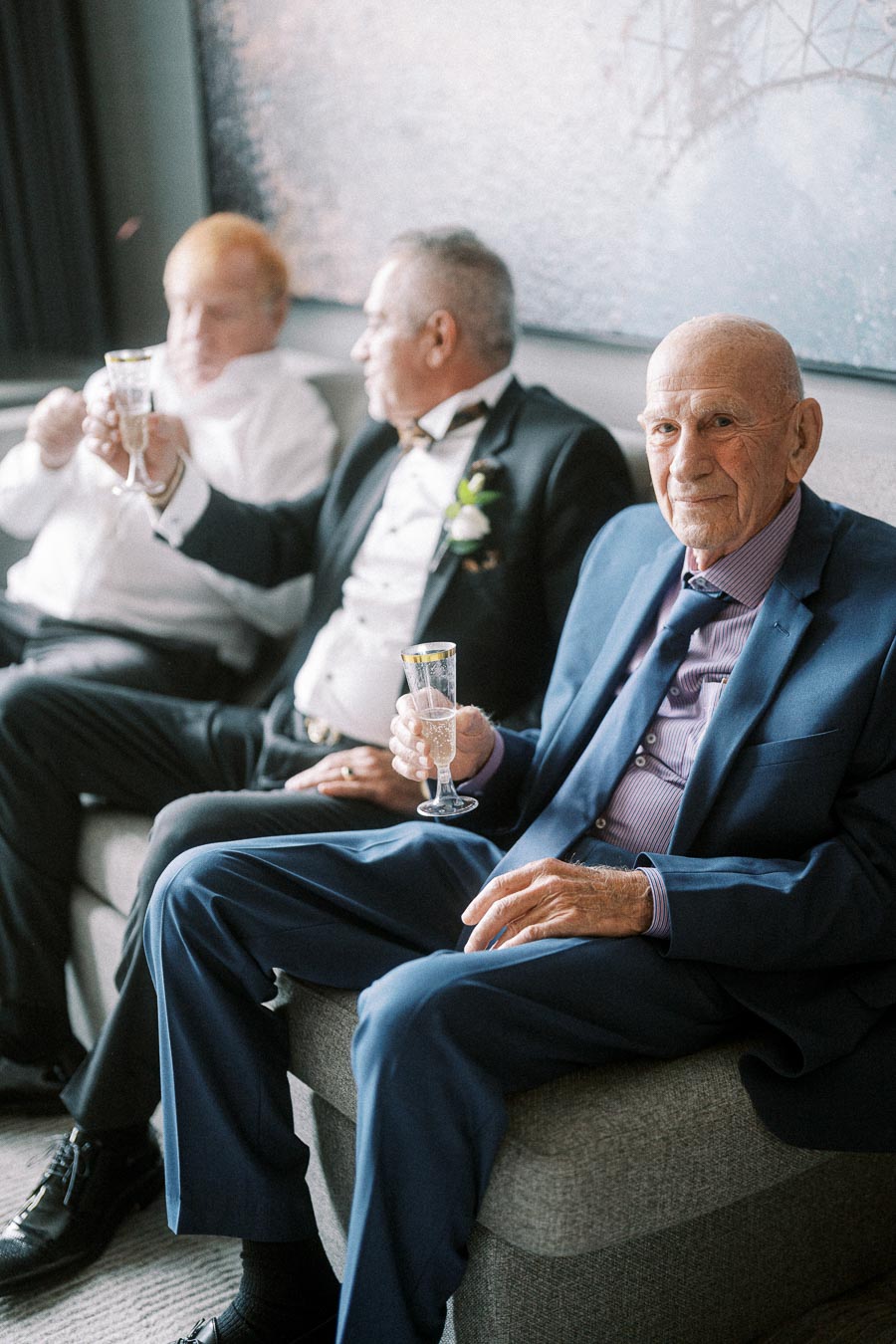 Elderly gentlemen in suits enjoying champagne while seated on a sofa, dressed for a formal occasion.